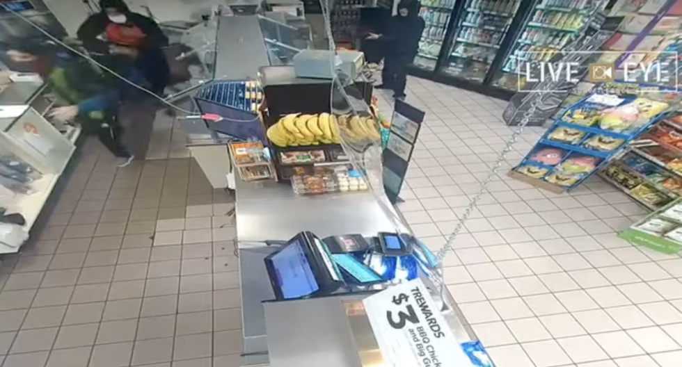 Robbers push a 7 Eleven clerk towards the cash register.