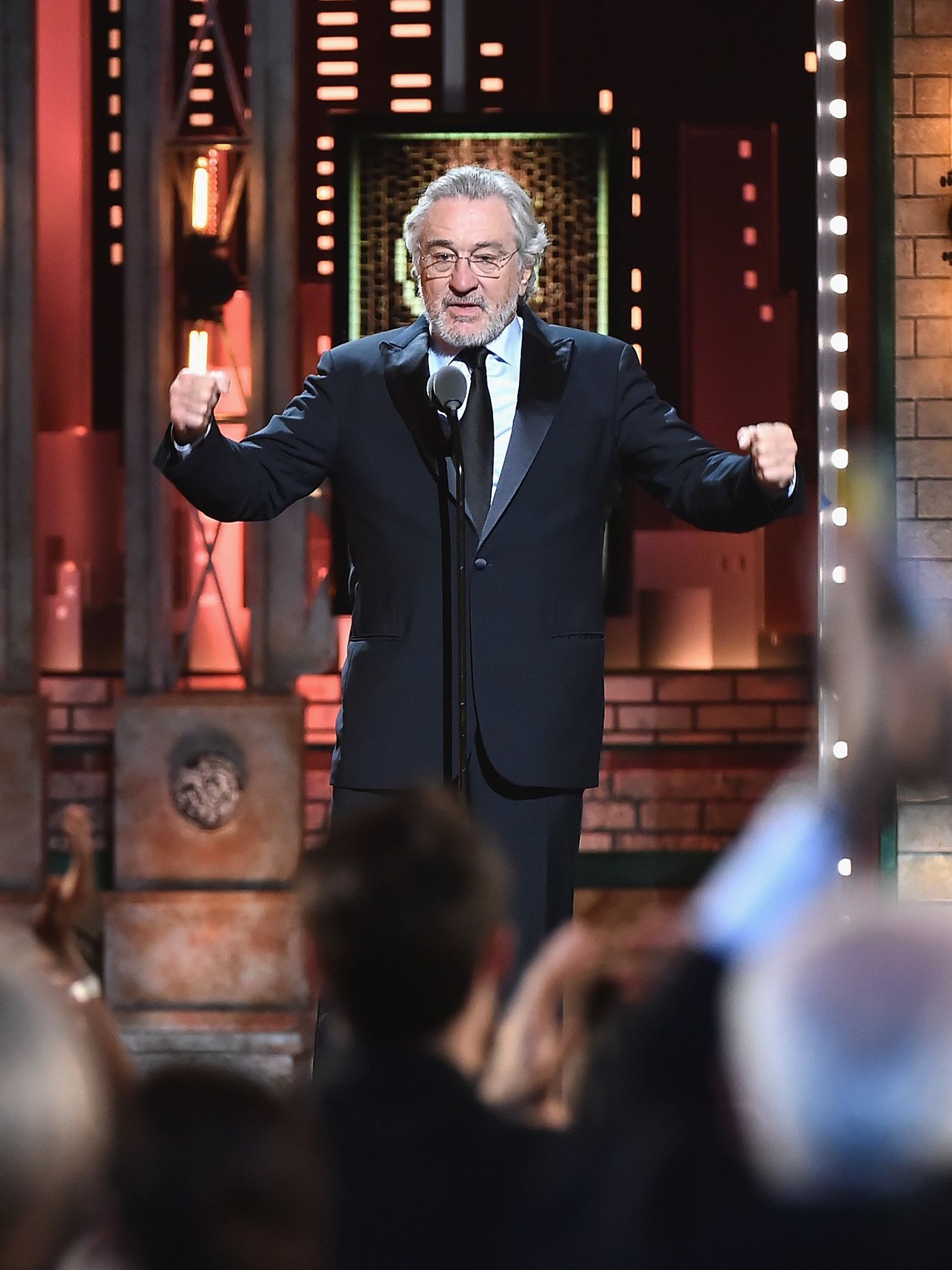 Robert De Niro, a bearded white man with short grey hair and a black suit, on stage at the Tony Awards in 2018.