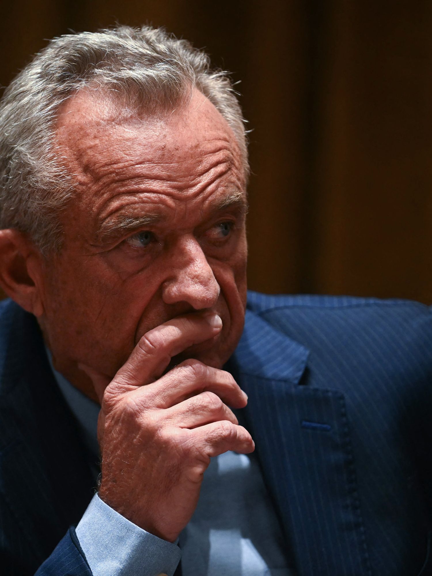 Robert F Kennedy Jr, a white man with short grey hair and a blue suit, places his right hand to his face, in thought.