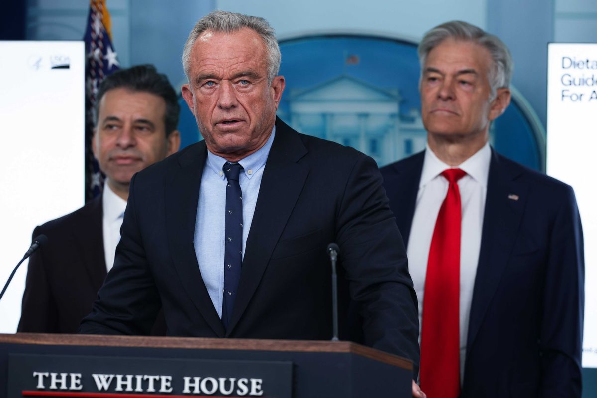 Robert F Kennedy Jr. stands behind the navy 'The White House' lectern wearing a black suit. Two black-suitedd man stsand heind RFK on either side.