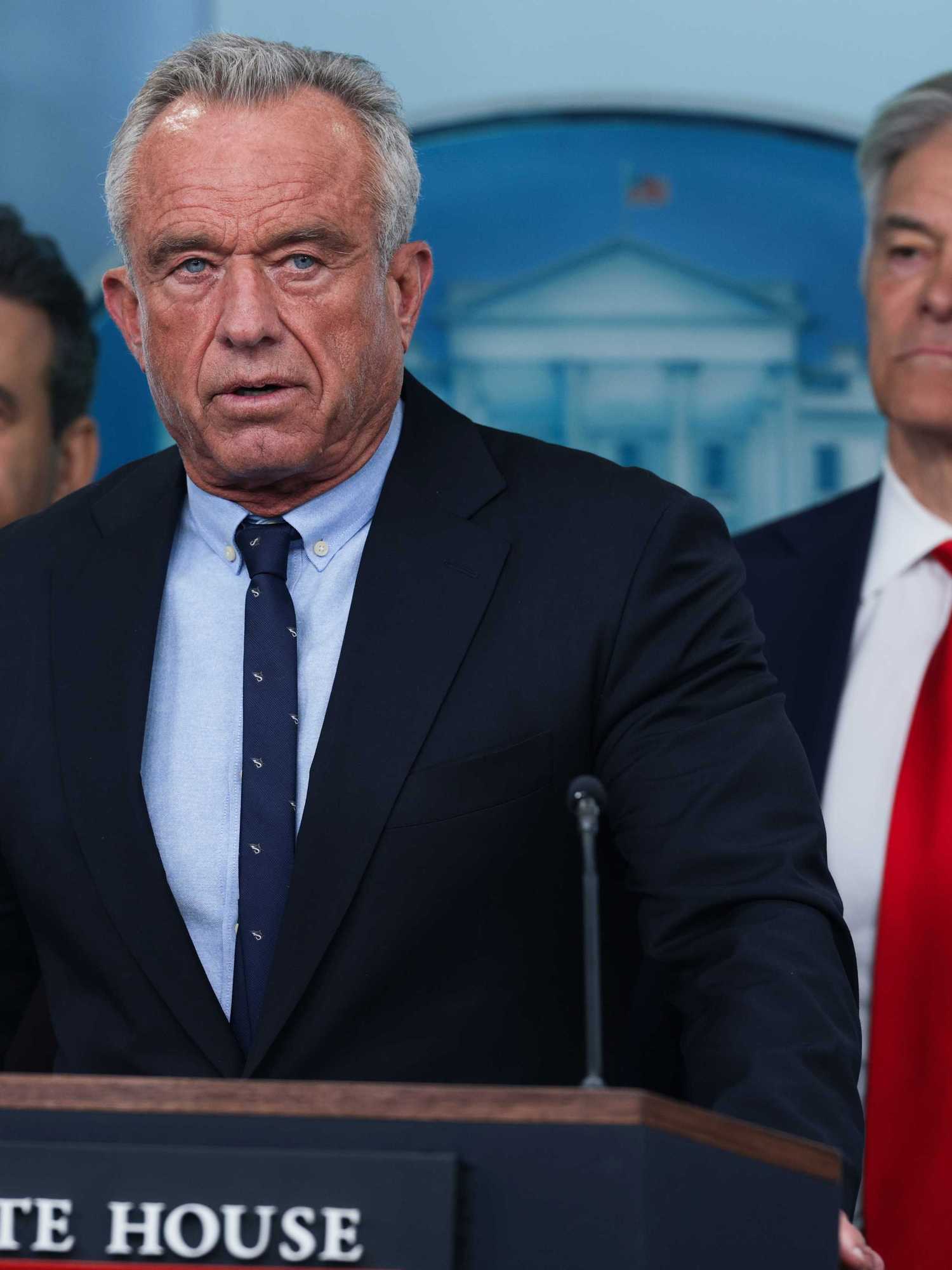 Robert F Kennedy Jr. stands behind the navy 'The White House' lectern wearing a black suit. Two black-suitedd man stsand heind RFK on either side.