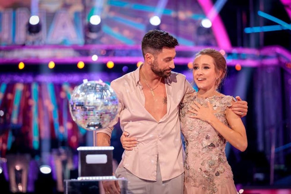 Rose Ayling-Ellis, a young white woman with short blonde hair, places her hand on her chest. She is embraced by Giovanni Pernice, a white man with short black hair, as they stand behind a glitter ball trophy on Strictly.