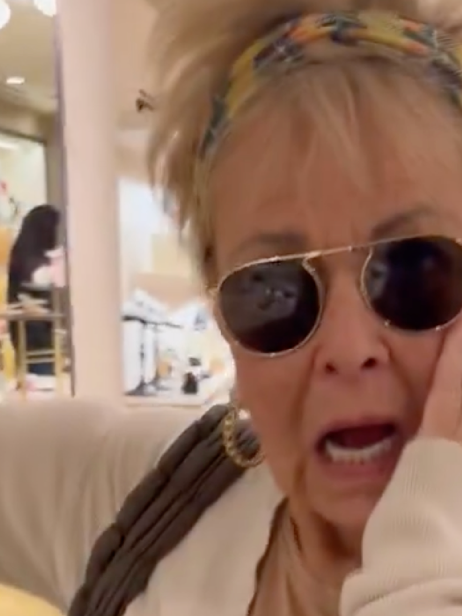 Roseanne Barr, a white woman with spiky blonde hair, black sunglasses and a white fleece, places her left hand to her face, looking shocked. She's in a department store.