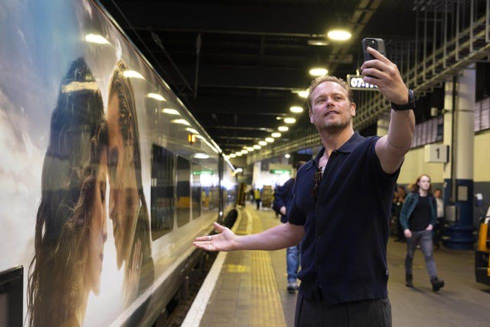 Sam Heughan takes a selfie of himself in front of a train featuring an Outlander-themed wrap