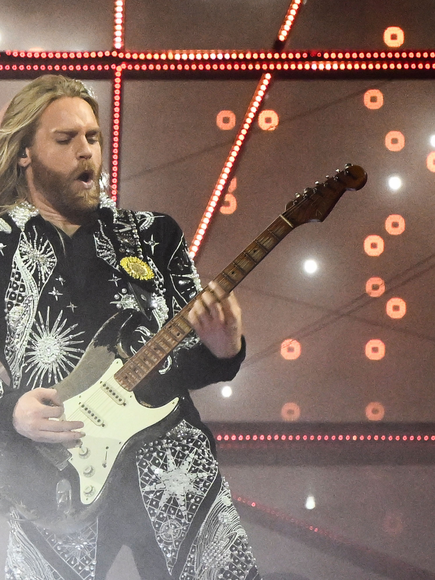 Sam Ryder, a bearded white man with long blonde hair, leans back slightly as he plays an electric guitar on stage at the Eurovision Song Contest.