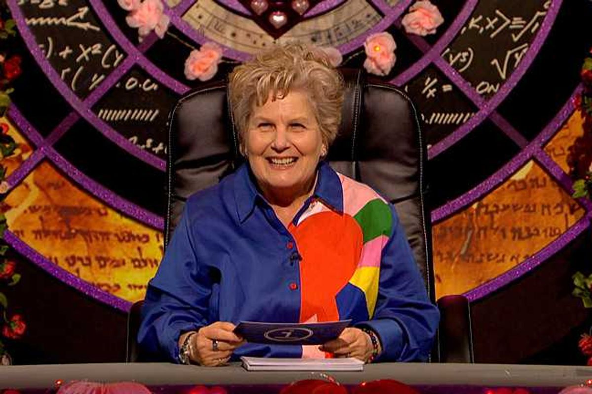 Sandi Toksvig, a white woman with short blonde hair and a blue shirt with colourful patterns on the left side, presenting an episode of QI.