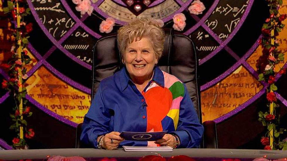Sandi Toksvig, a white woman with short blonde hair and a blue shirt with colourful patterns on the left side, presenting an episode of QI.