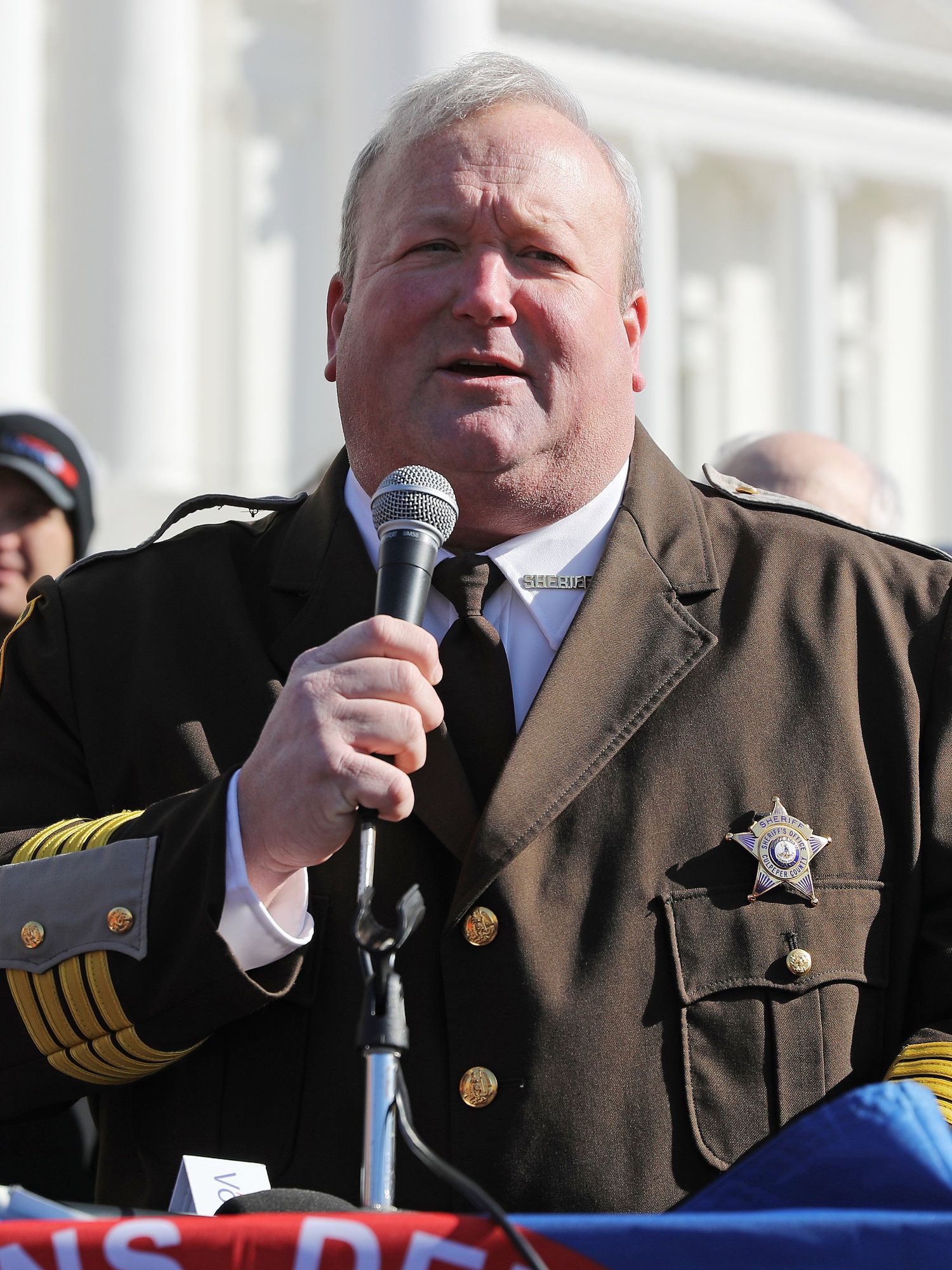 Scott Jenkins, a white man with short grey hair and a brown sheriff uniform, speaking into a microphone at a rally.