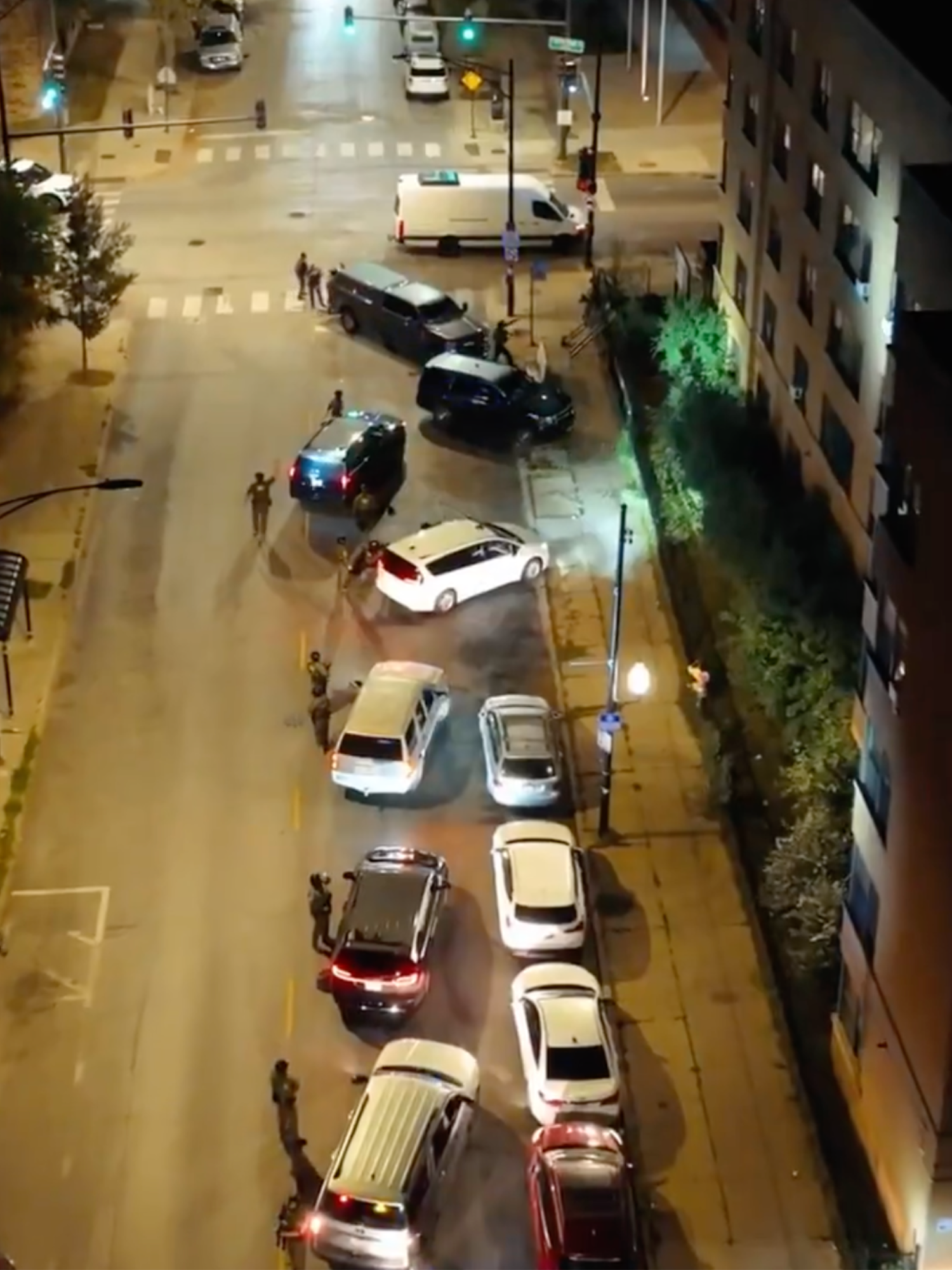 Several white cars and vans are parked outside an apartment building at night.