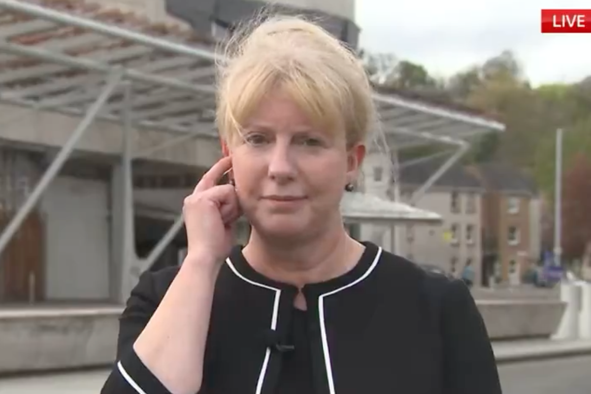 Shona Robison, a white woman with short blonde hair and a black dress, places a finger to her ear to hear an earpiece during a Sky News interview in front of Scottish parlament.
