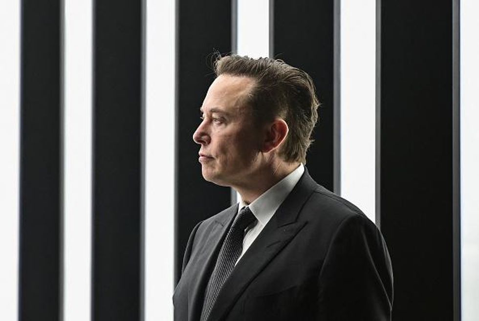 Side profile of Elon Musk, a white man with short black hair and a black suit, standing in front of some white beams of light at a Tesla event.