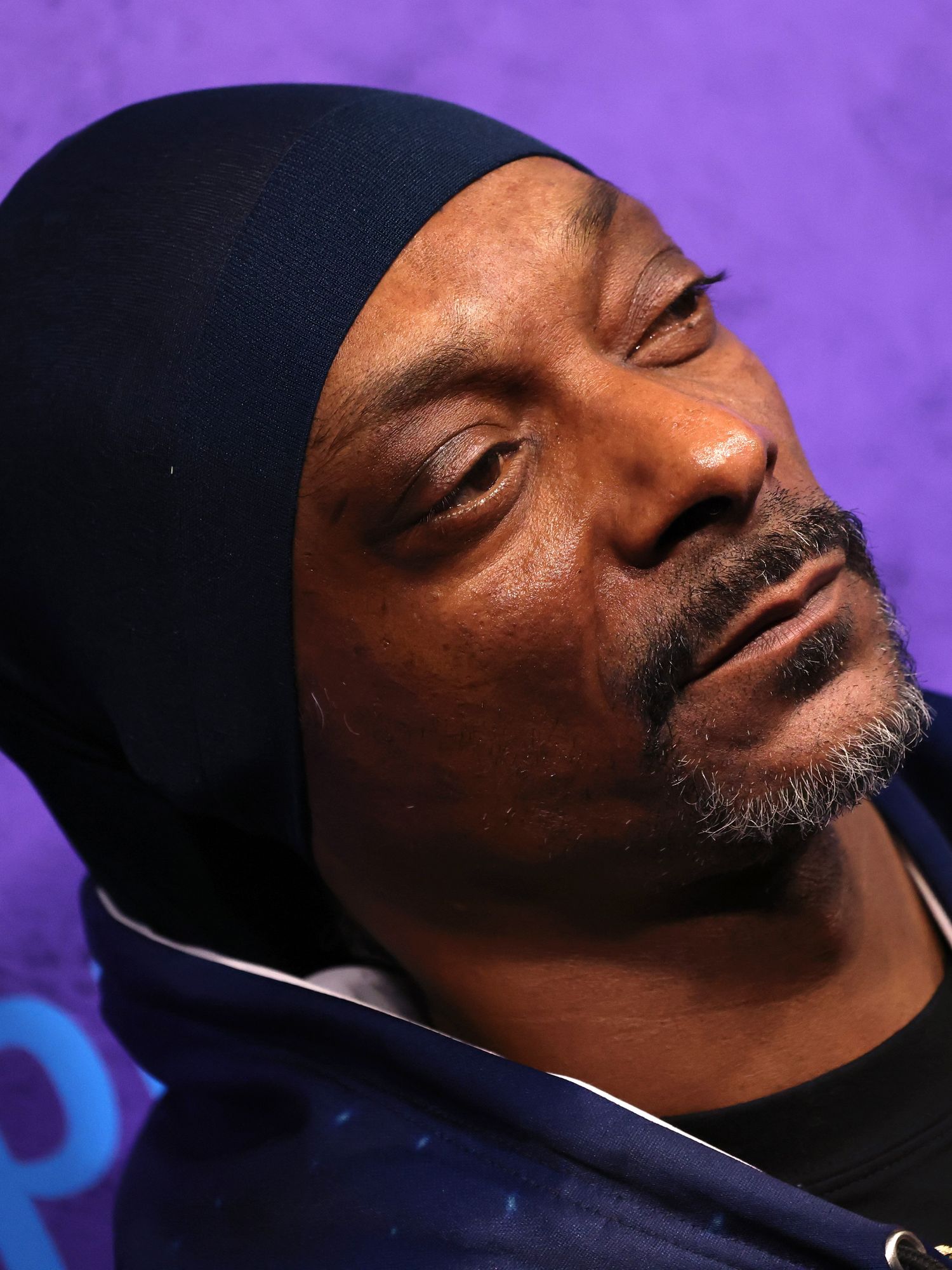 Side profile of Snoop Dogg, a Black man with a black durag, black beard and blue hoodie against a purple posterboard.