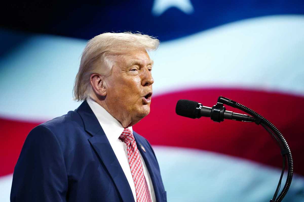 Side shot of Donald Trump speaking into a microphone with a US flag on a screen behind him.