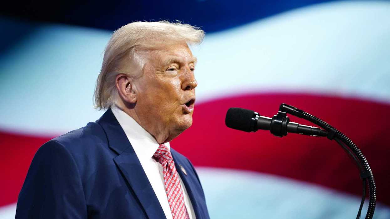Side shot of Donald Trump speaking into a microphone with a US flag on a screen behind him.