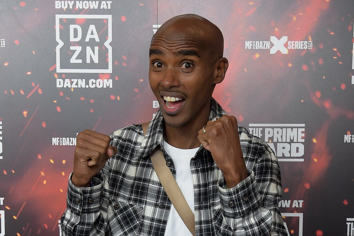 Sir Mo Farah smiles and raises his fists in front of a DAZN posterboard.