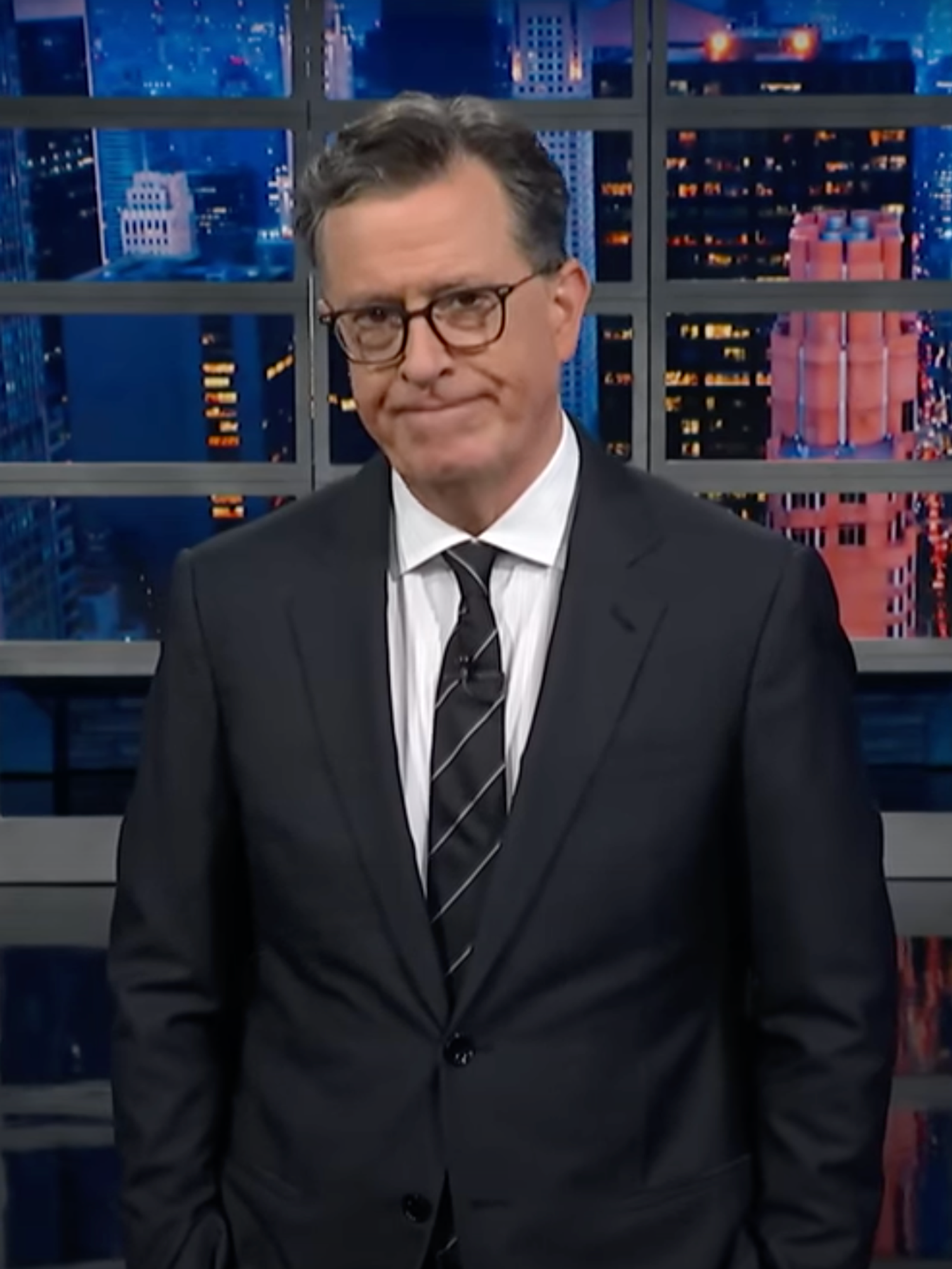 Stephen Colbert, a white man with short black hair and glasses, wearing a black suit and tie and looking disappointed as he presents his TV show.