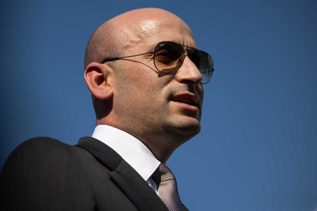 Stephen Miller, a bald white man in a black suit with brown tinted sunglasses.