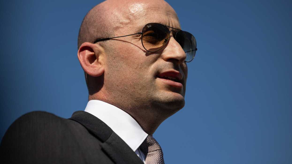 Stephen Miller, a bald white man in a black suit with brown tinted sunglasses.