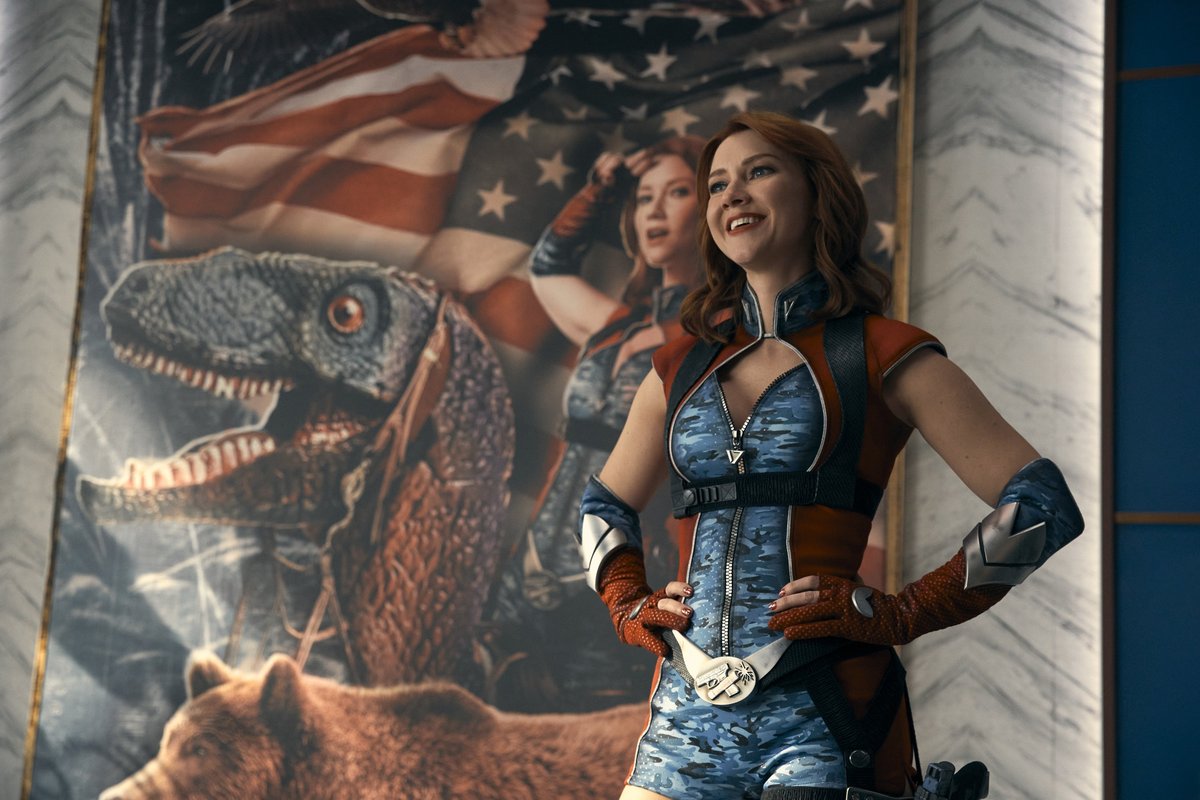 Still from The Boys. Firecracker, a white woman with wavy ginger hair and a blue and red superhero suit, smiles with her hands on her hips in front of a giant poster with a dinosaur and American flag.