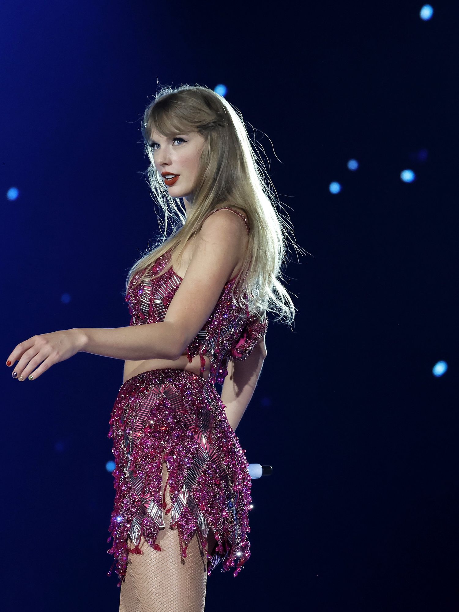Taylor Swift, a white woman with long blonde hair in a glittery pink dress, stands on stage during her Eras tour.