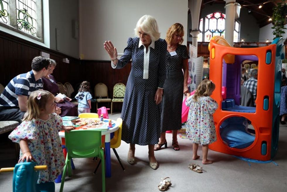 The Queen waves to a child near a toy table while other children play watched by adults