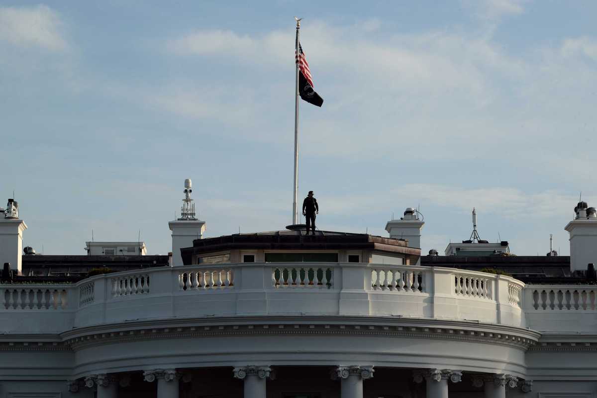 The top of the White House building, where an American flag is erected on a flag pole and a man is standing at the bottom of it.