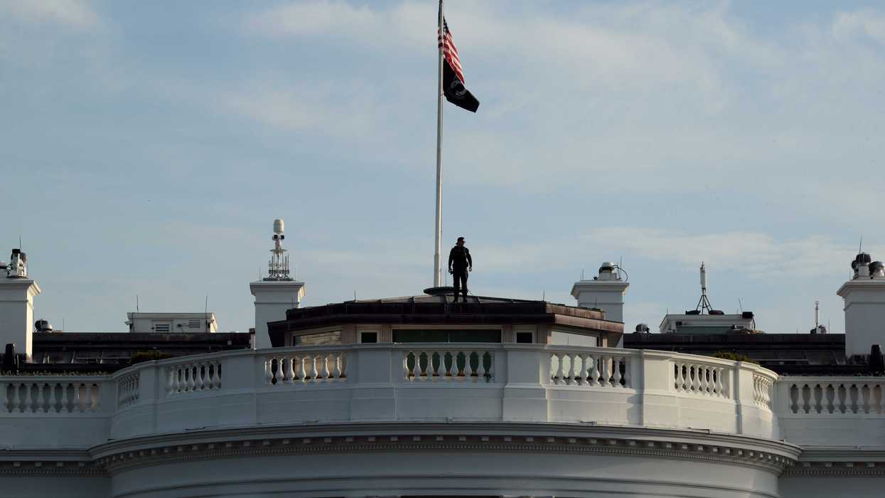 The top of the White House building, where an American flag is erected on a flag pole and a man is standing at the bottom of it.