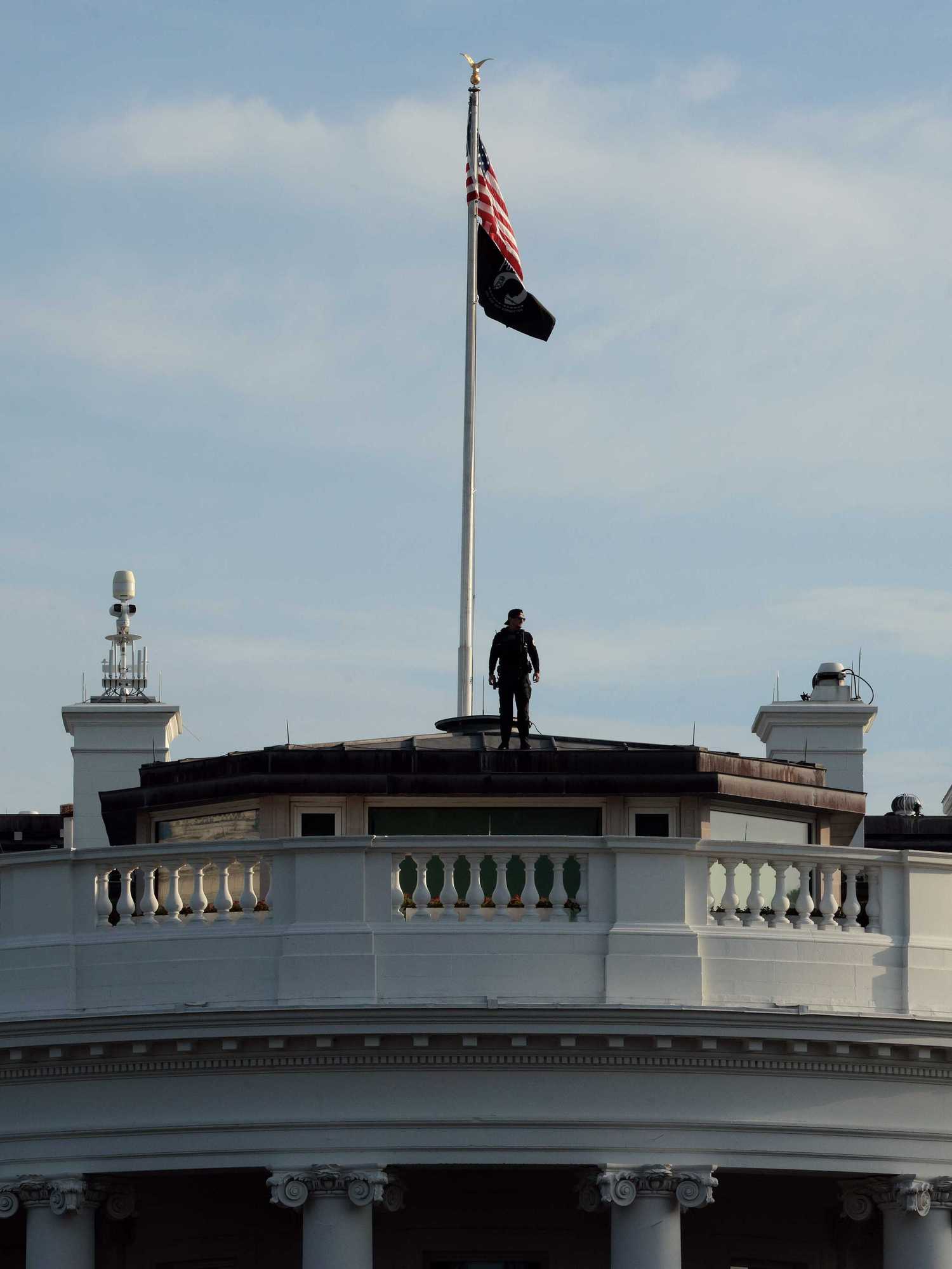 The top of the White House building, where an American flag is erected on a flag pole and a man is standing at the bottom of it.