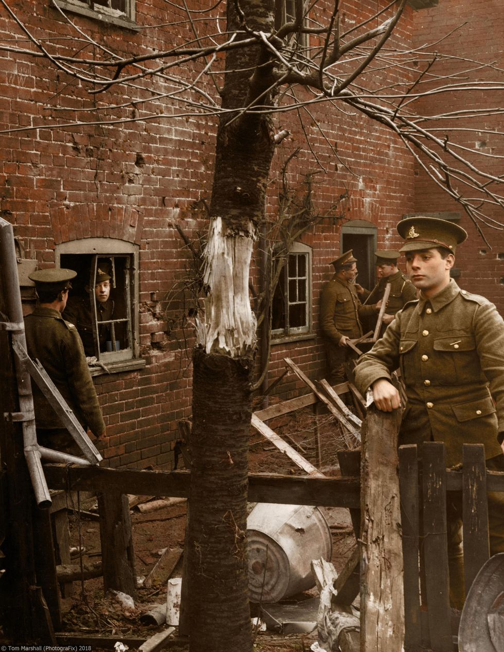 This photograph shows the aftermath of a Zeppelin raid, which has partially stripped the bark off a tree and blown out windows in a building. First World War soldiers are seen inspecting the area in the aftermath of the blast.