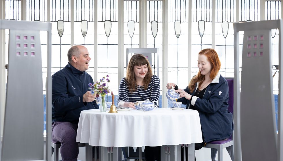 Three people sat in a row at a round table covered by a white tablecloth, looking happy, with one pouring tea from an ornate teapot into a cup