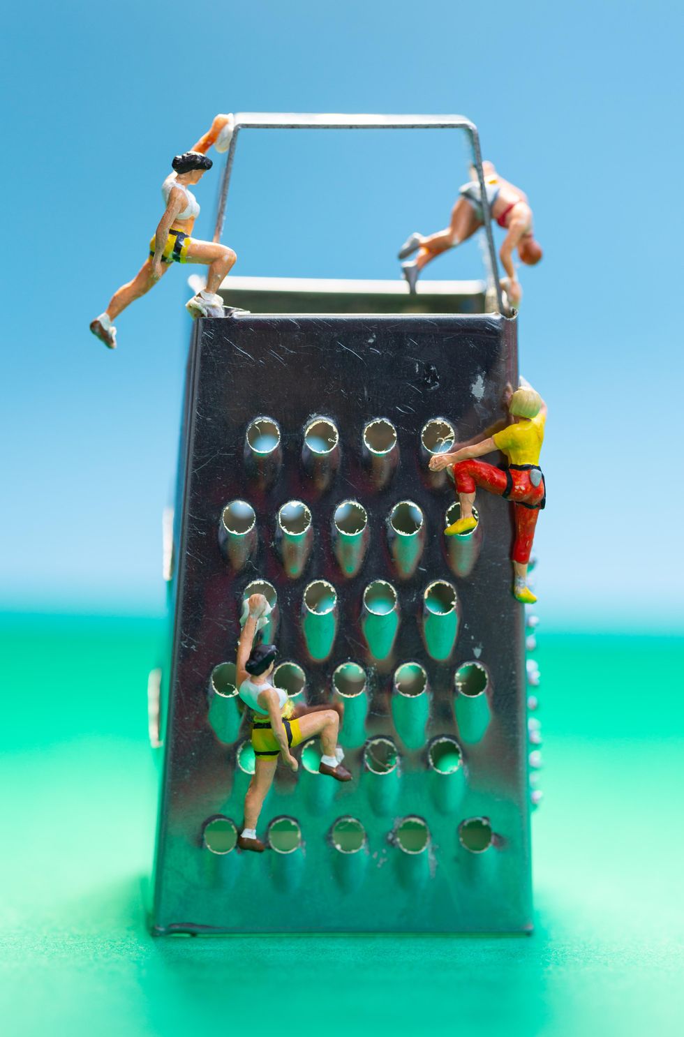Tiny figures on a cheese grater, posed as if using it as a climbing wall