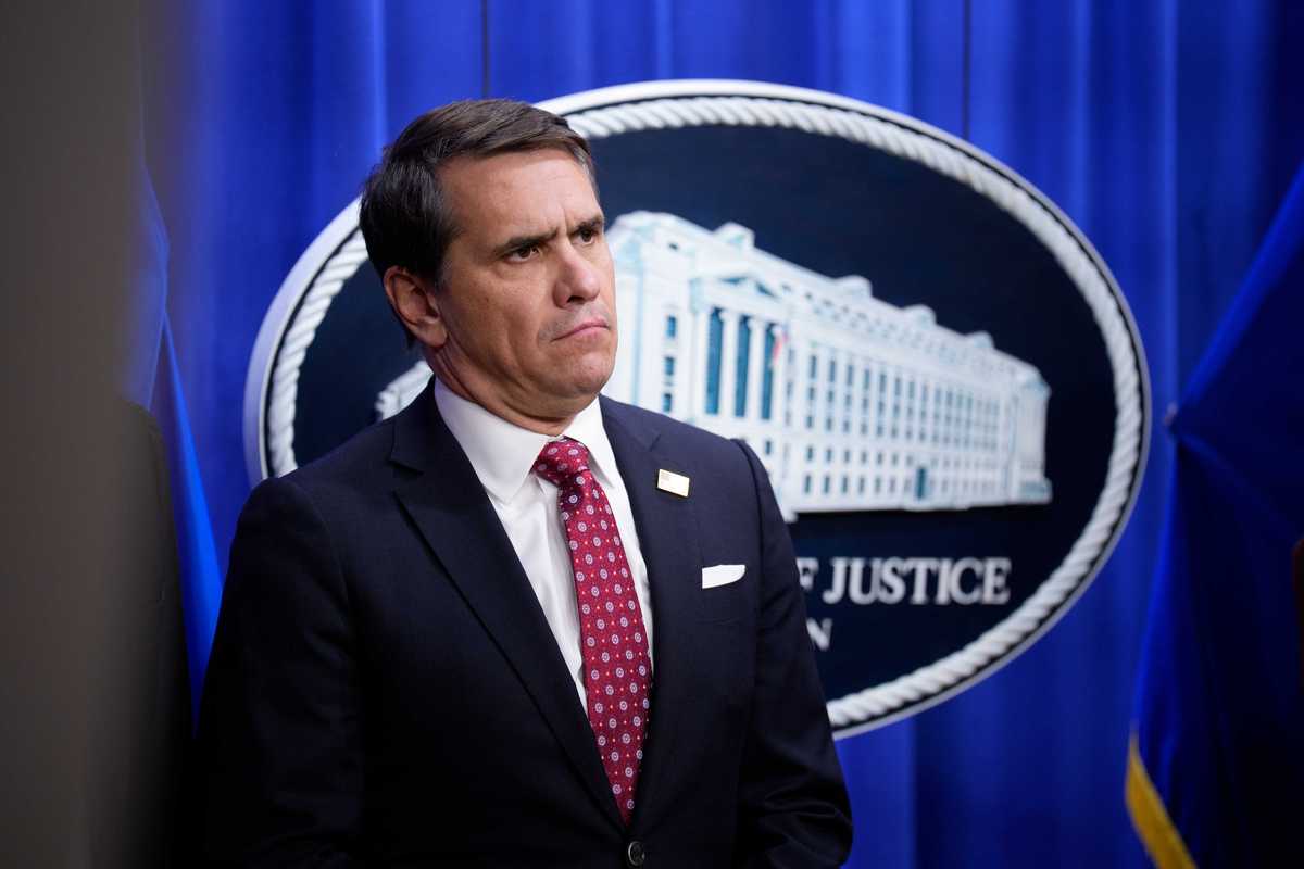 Todd Blanche, a white man with short black hair, a black suit and a red tie, standing in front of a blue Department of Justice curtain.