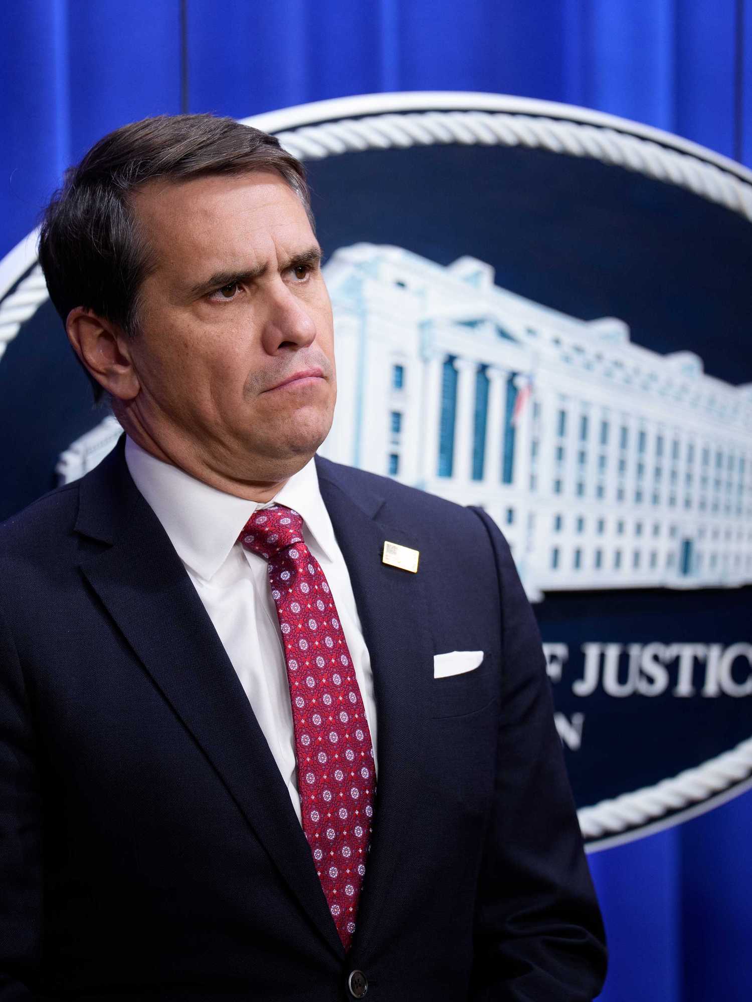 Todd Blanche, a white man with short black hair, a black suit and a red tie, standing in front of a blue Department of Justice curtain.