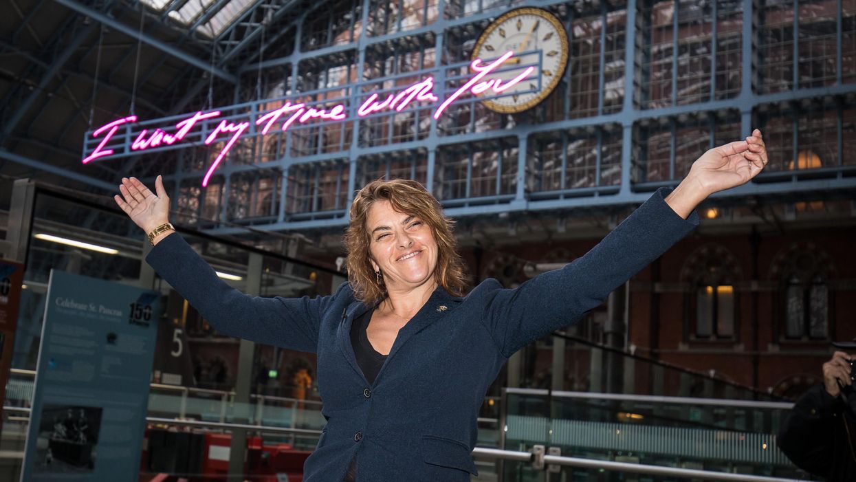 Tracey Emin wearing a blue jacket holds her arms up in a Y shape in front of a neon sign hanging up in a train station behind her. The handwritten text reads: 'I want my time with you'.