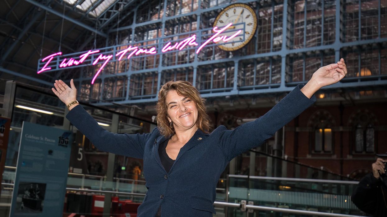 Tracey Emin wearing a blue jacket holds her arms up in a Y shape in front of a neon sign hanging up in a train station behind her. The handwritten text reads: 'I want my time with you'.