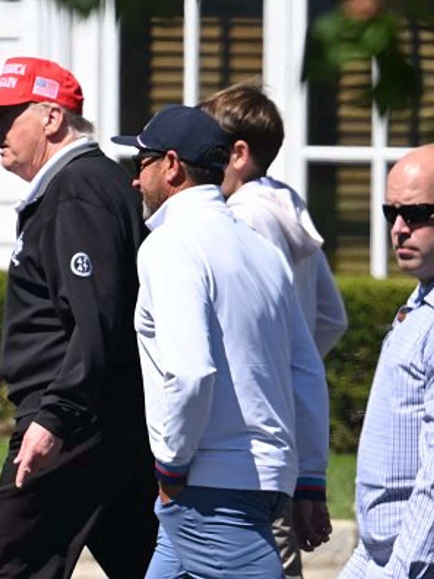 Trump, in a black jacket and red MAGA cap, walks around his golf course with his security detail.