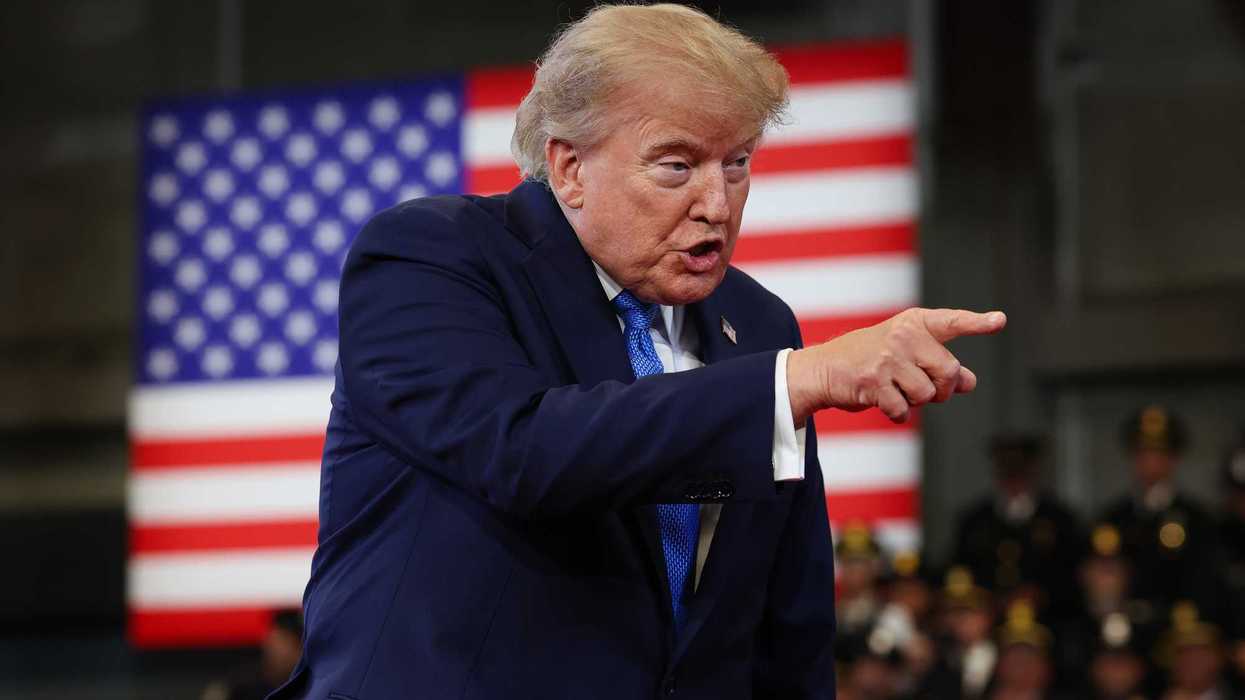 Trump points with his right hand, on stage, at someone off-camera. An American flag is visible hanging behind him.