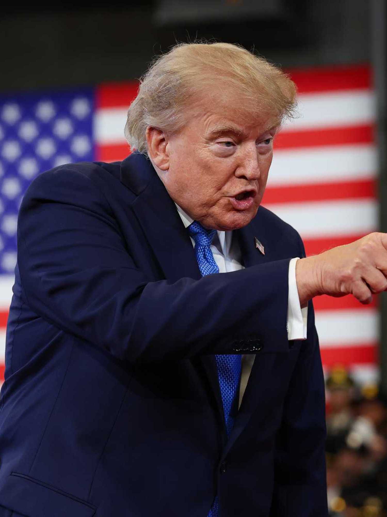 Trump points with his right hand, on stage, at someone off-camera. An American flag is visible hanging behind him.