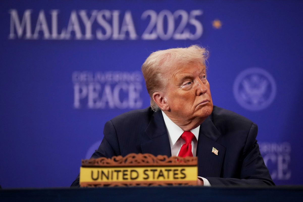 US president Donald Trump in a black suit with a red tie, sitting at a table with a text sign on it reading 'United States'. He's looking to his left with a neutral expression on his face. Out of focus in the background is a blue poster board with the words 'Malaysia 2025' on it.