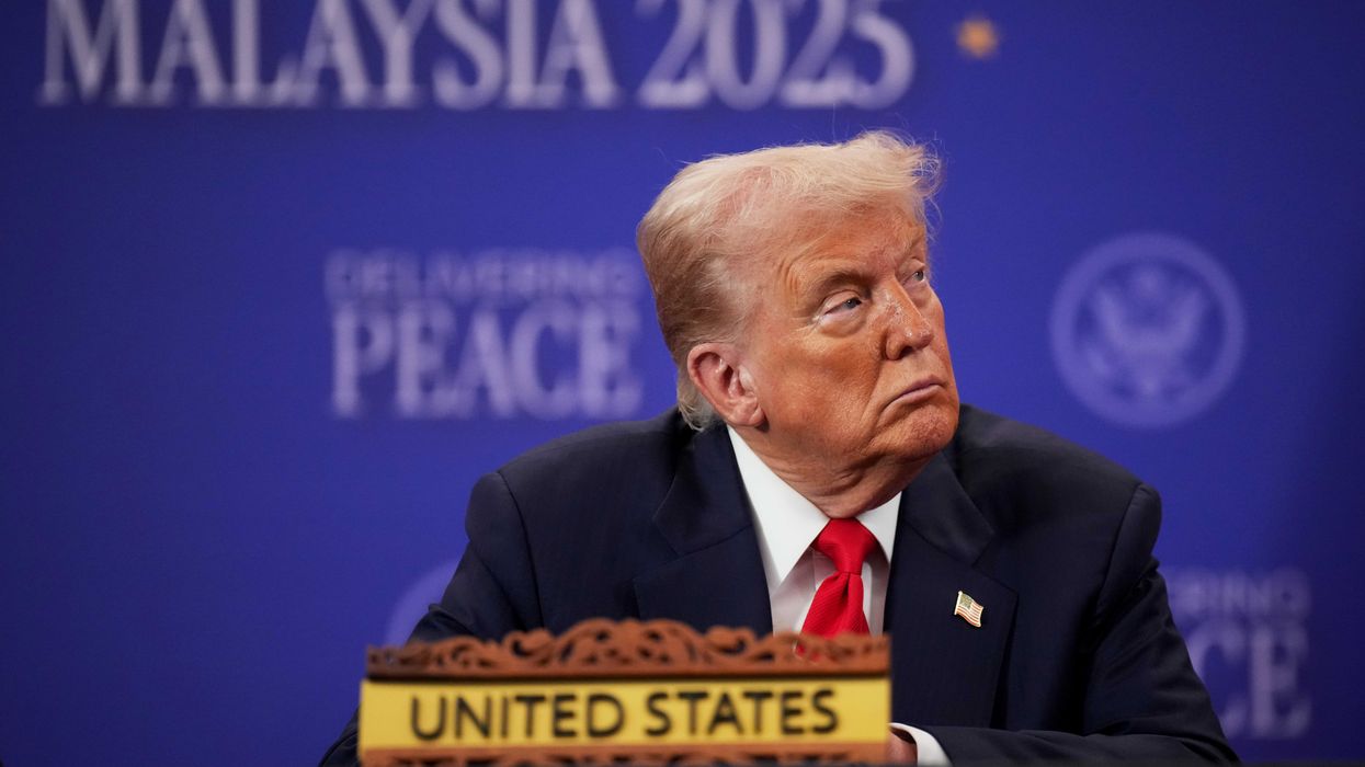 US president Donald Trump in a black suit with a red tie, sitting at a table with a text sign on it reading 'United States'. He's looking to his left with a neutral expression on his face. Out of focus in the background is a blue poster board with the words 'Malaysia 2025' on it.