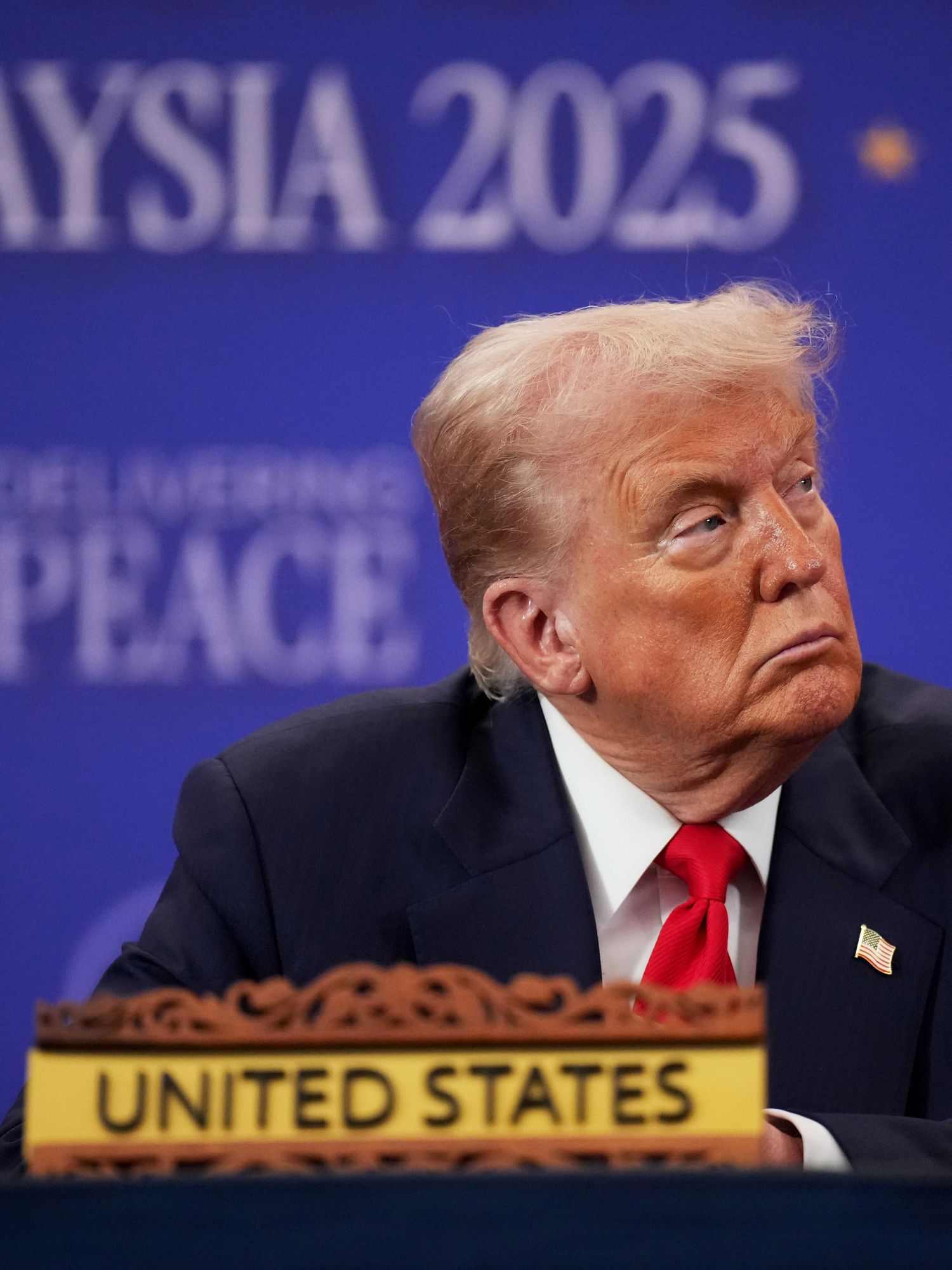 US president Donald Trump in a black suit with a red tie, sitting at a table with a text sign on it reading 'United States'. He's looking to his left with a neutral expression on his face. Out of focus in the background is a blue poster board with the words 'Malaysia 2025' on it.