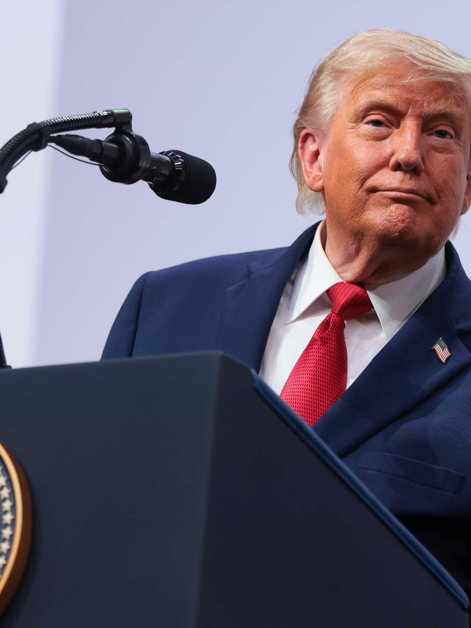 US president Donald Trump in a navy suit and red tie stands behind his presidential lectern on stage.