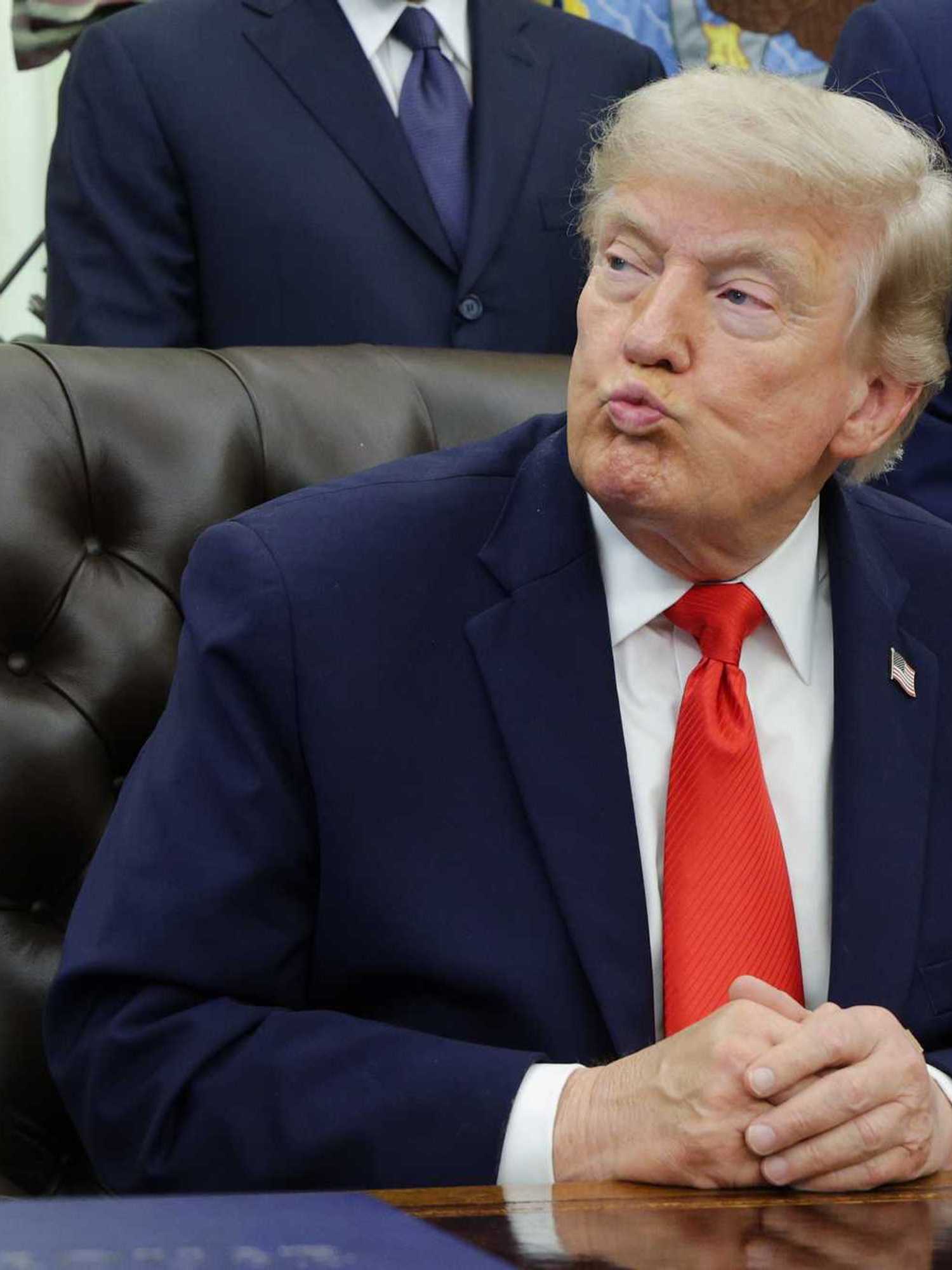 US president Donald Trump sits behind the Resolute Desk in the Oval Office, looking to his right and pursing his lips.