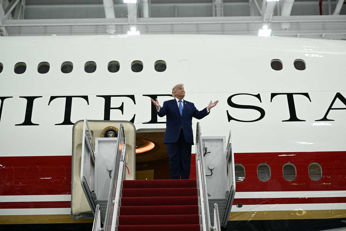 US president Donald Trump standing outside the new Air Force One plane, gesturing with his hands lifted upwards.