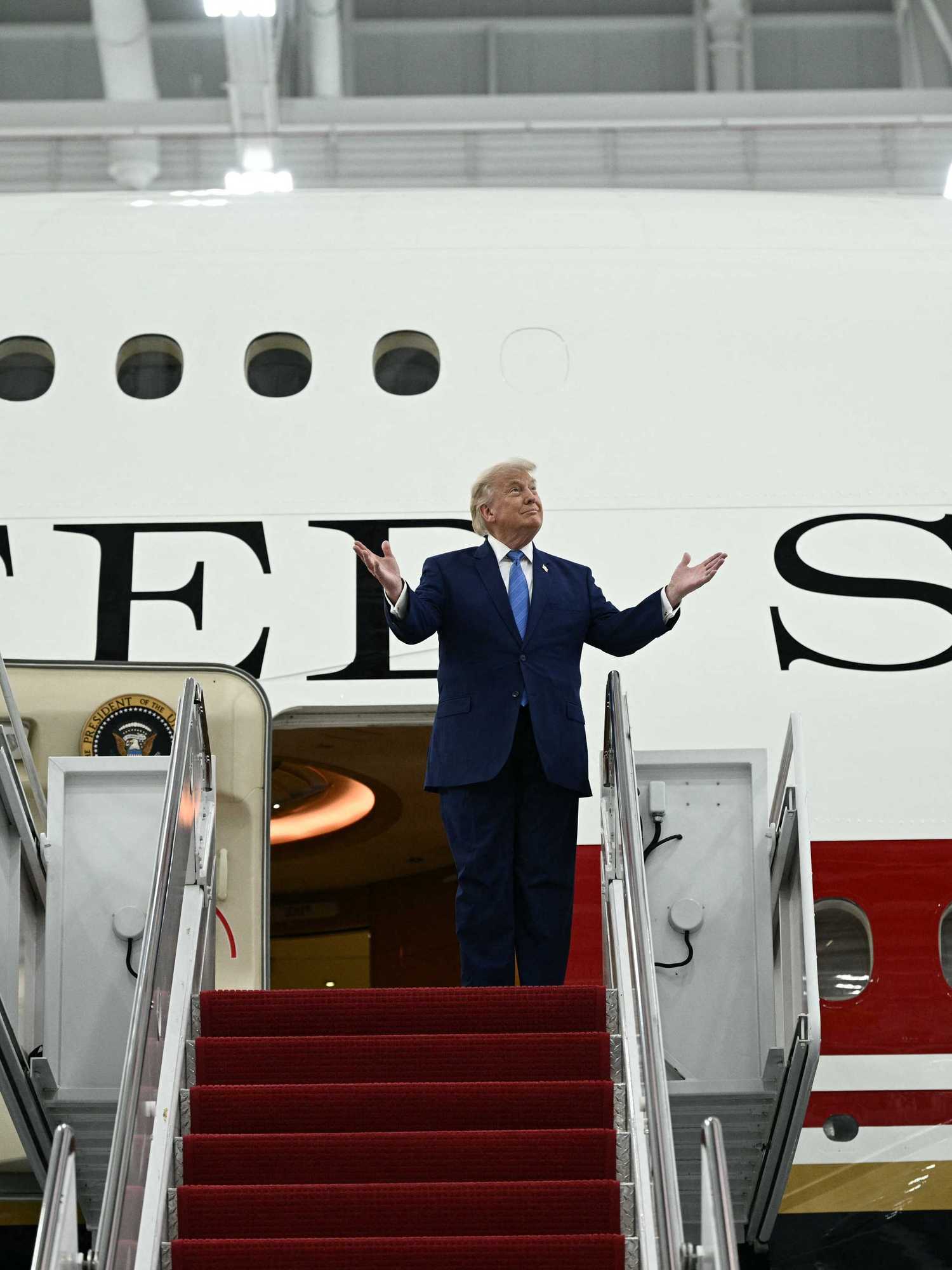 US president Donald Trump standing outside the new Air Force One plane, gesturing with his hands lifted upwards.