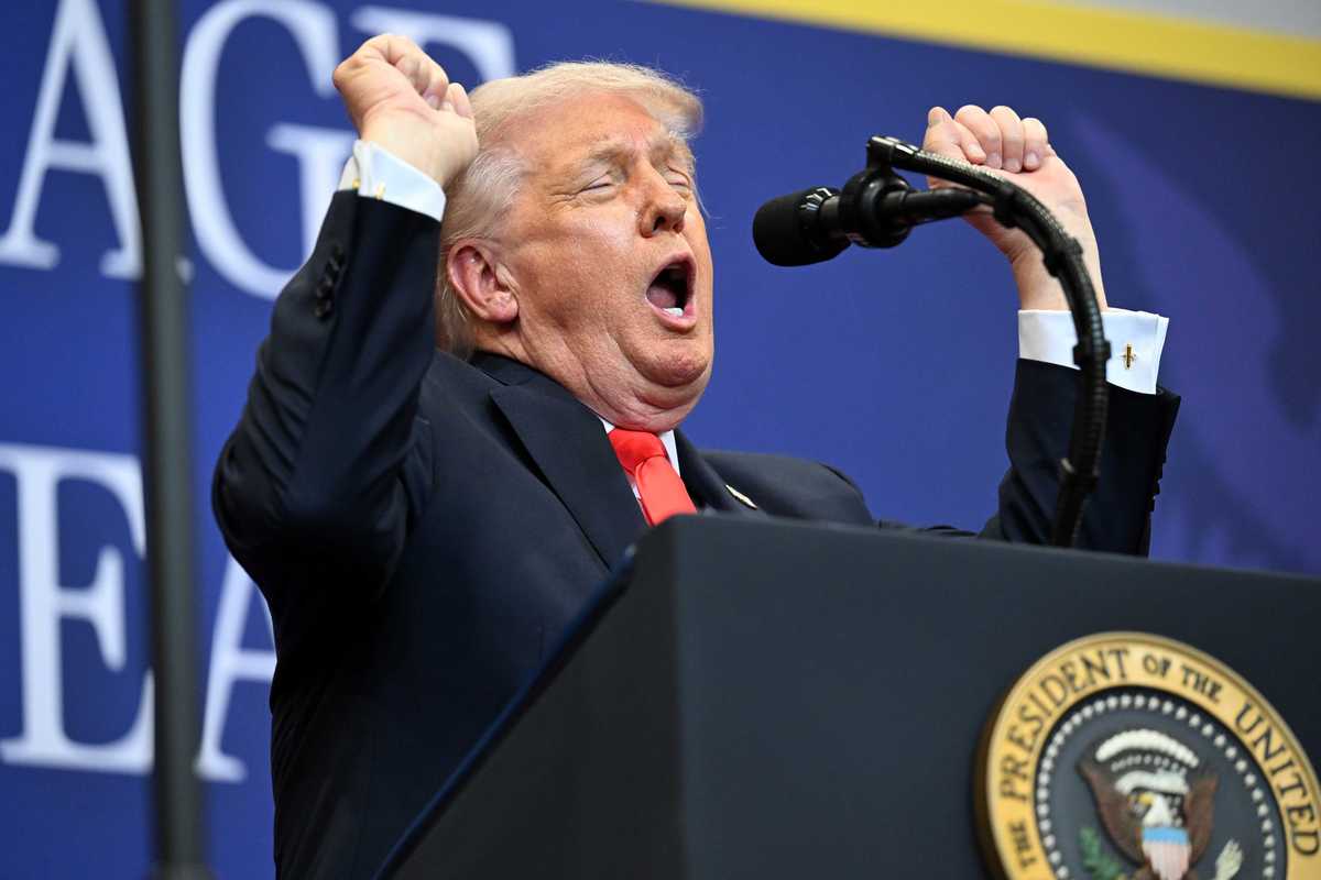US president Donald Trump stands behind the presidential lectern, raising both of his arms in a fist, closing his eyes and opening his mouth. He's impersonating a weightlifter.