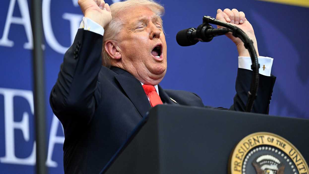 US president Donald Trump stands behind the presidential lectern, raising both of his arms in a fist, closing his eyes and opening his mouth. He's impersonating a weightlifter.