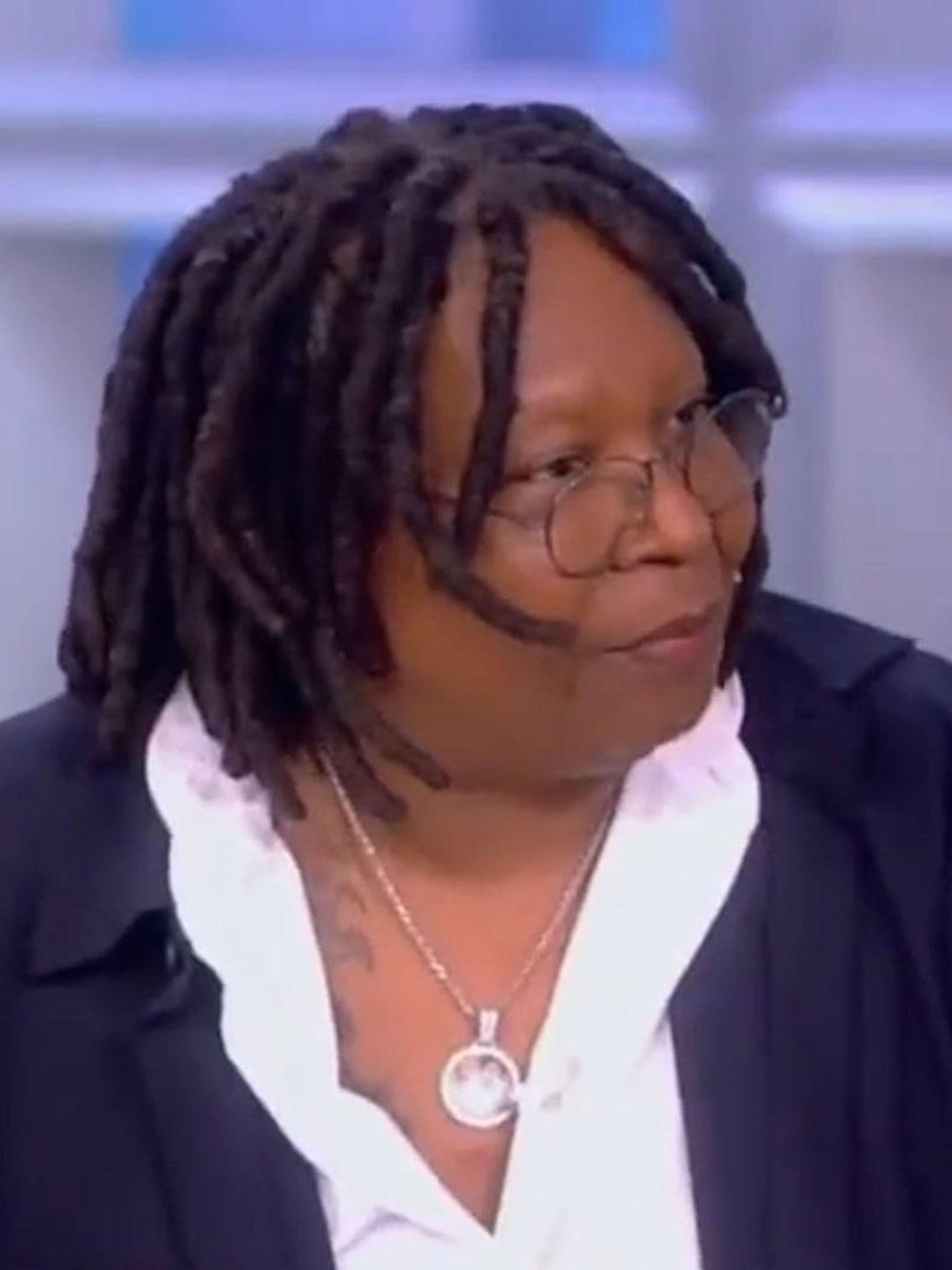 Whoopi Goldberg, a black woman with black dreadlocks sits on a panel, wearing a blue jacket and white shirt.