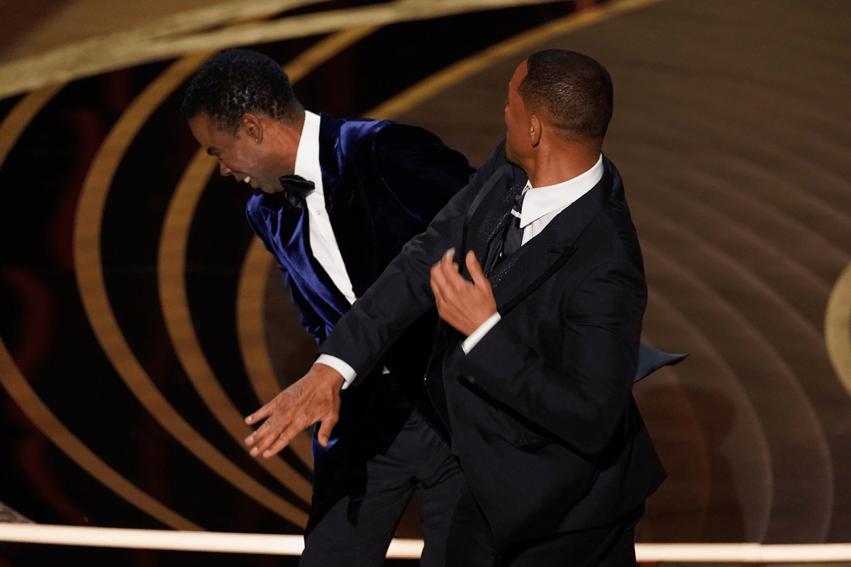 Will Smith, a Black man with short black hair and a black suit, slaps Chris Rock, a Black man with short black hair in a blue suit, on stage at the Oscars.