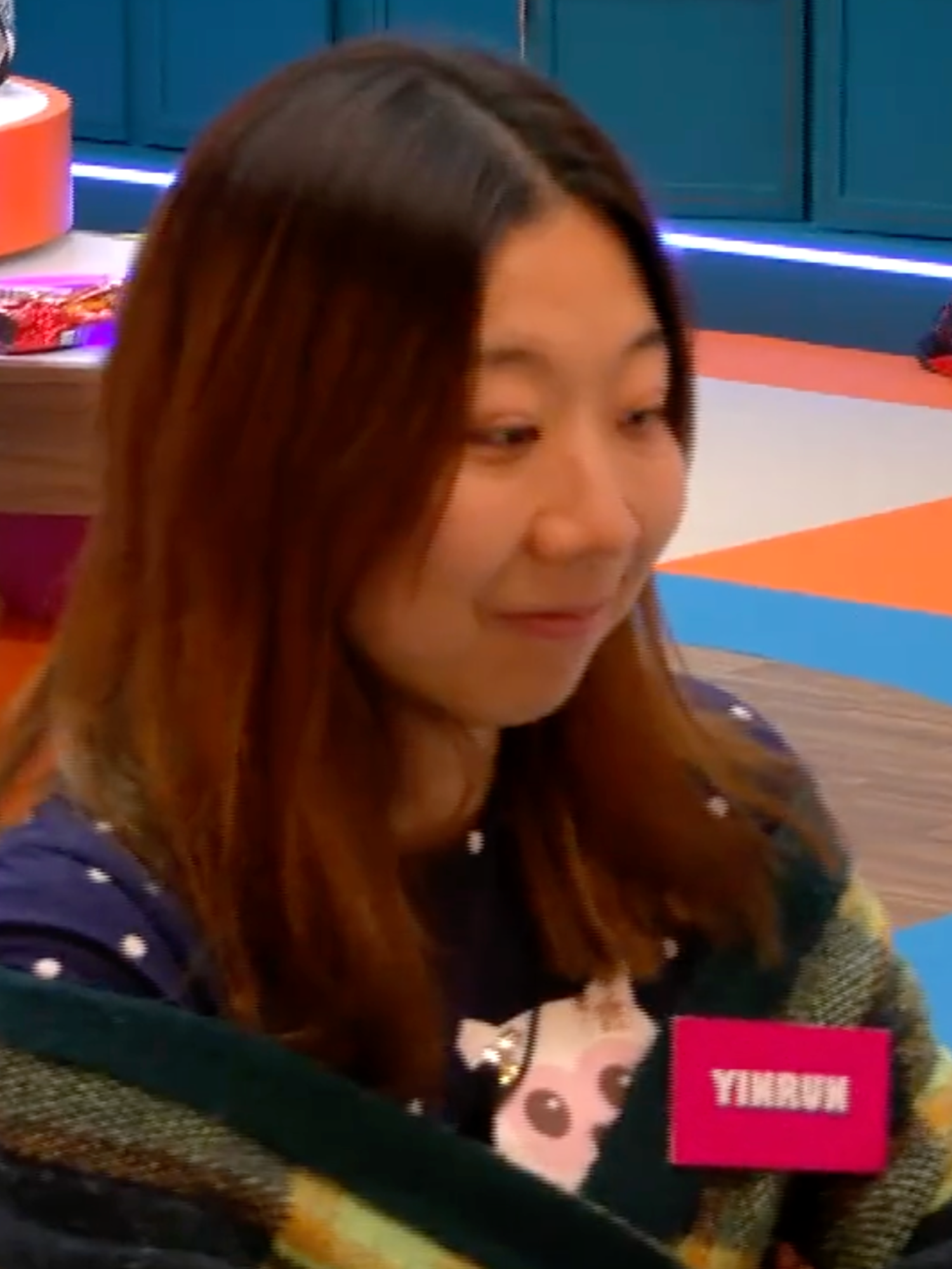 Yinrun, a young Chinese woman with long brown hair and a spotted blue shirt, wraps a green cardigan around herself and smiles while looking at someone in front of her.